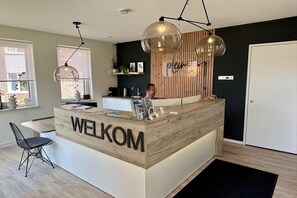 A reception desk with 'WELKOM' signage, a modern pendant light, a wooden accent wall, and a door to the right. - Ground floor lodge with private veranda (Zoutelande)