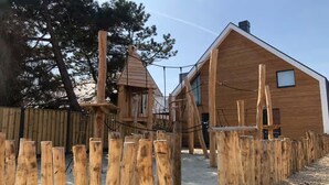 Une cabane en bois avec un toit en pente et une cheminée, entourée d’une clôture en bois et d’une structure de jeux pour enfants.