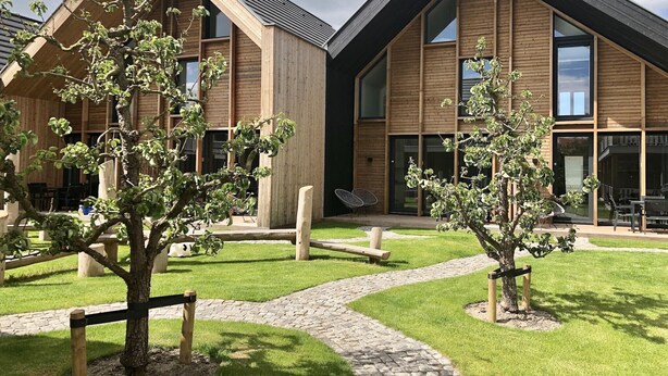 Een modern gebouw met een houten gevel, grote ramen en een bestraat erf met bomen en zitplaatsen.