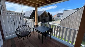 A covered balcony with a hanging chair, a table, and a view of neighboring buildings. - Lodge with beautiful view of garden (Zoutelande)
