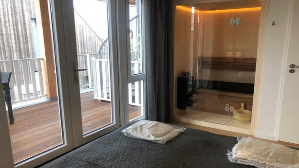 A room with a large sliding glass door leading to a wooden deck, a bed with a gray quilt, and a small table with a white pillow. - Lodge with beautiful view of garden (Zoutelande)