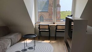 A room with a sofa, a small round table, two chairs, a TV, and a window with a view of a brick building. - 2-person lodge on 2nd floor with view of dunes. (Zoutelande)