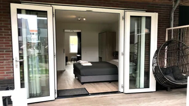 A modern bedroom with a large bed, a hanging chair, and a sliding glass door leading to a bathroom. - Lodge with beautiful view of the church and dunes. (Zoutelande)
