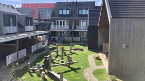 A courtyard with a playground, a building with a balcony, and a red-roofed structure in the background. - Lodge with beautiful view of the church and dunes. (Zoutelande)