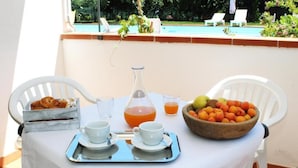 Une table dressée pour le petit-déjeuner, avec des croissants, du jus de fruits et un saladier de fruits, entourée de chaises blanches, et donnant sur une piscine.