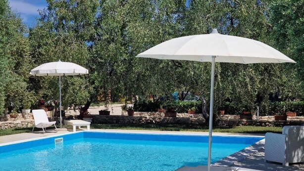 Une piscine entourée de verdure et de murs en pierre, avec des chaises longues blanches et des parasols.