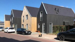 Una hilera de casas modernas con techos oscuros y fachadas de madera, coches aparcados y un cielo azul despejado.