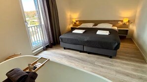 Een hotelkamer met een groot bed, twee nachtkastjes met lampen, een badkuip en een schuifdeur naar het balkon.