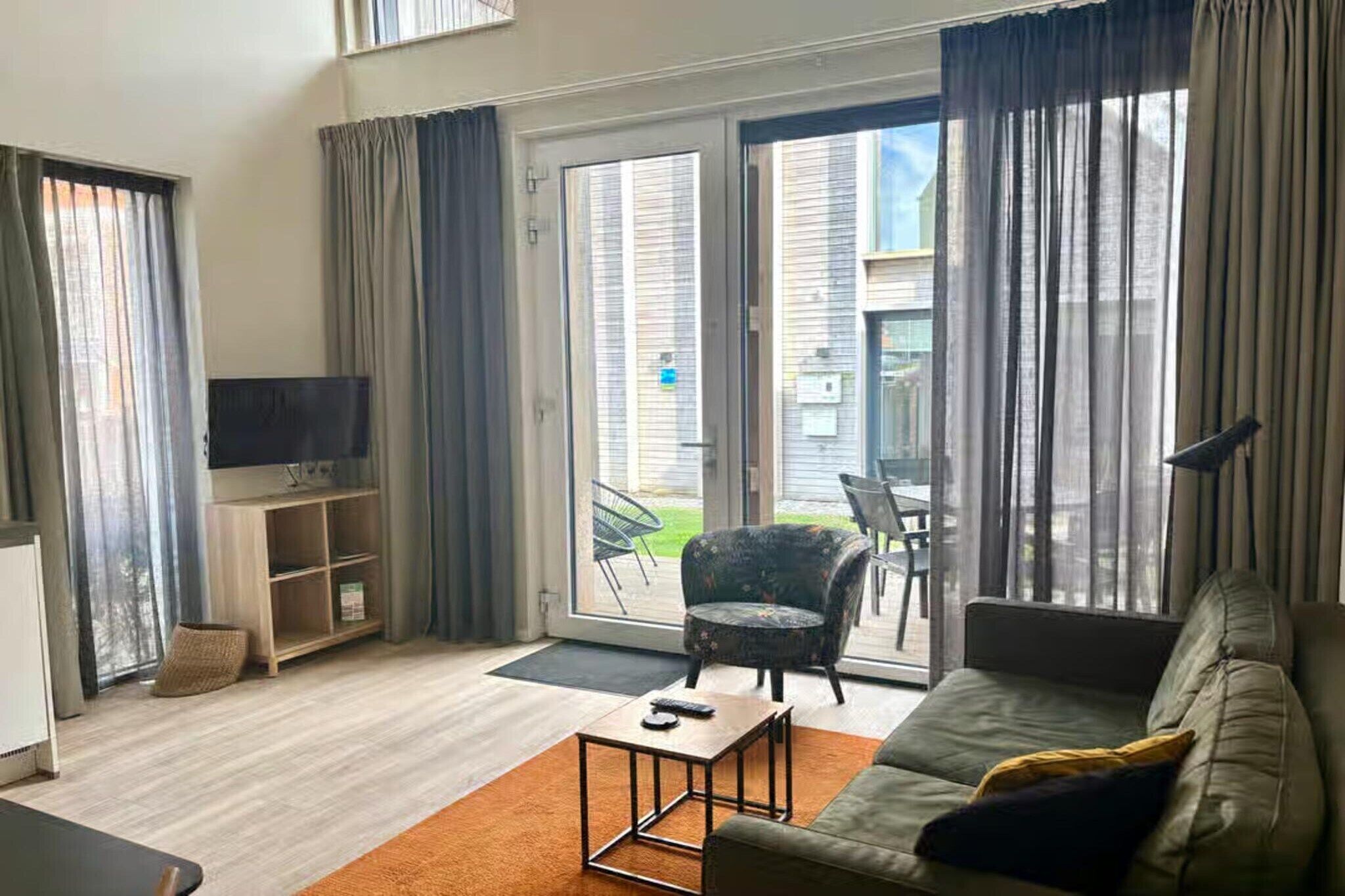 Uma sala de estar moderna com sofá, mesa de centro, televisão e porta de vidro deslizante que dá para uma área externa com uma cadeira e mesa.