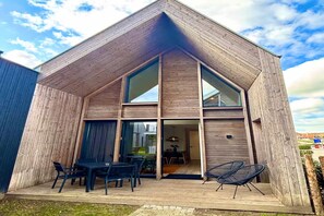 Een modern houten huis met een schuin dak, grote ramen en een open terras met buitenmeubilair.