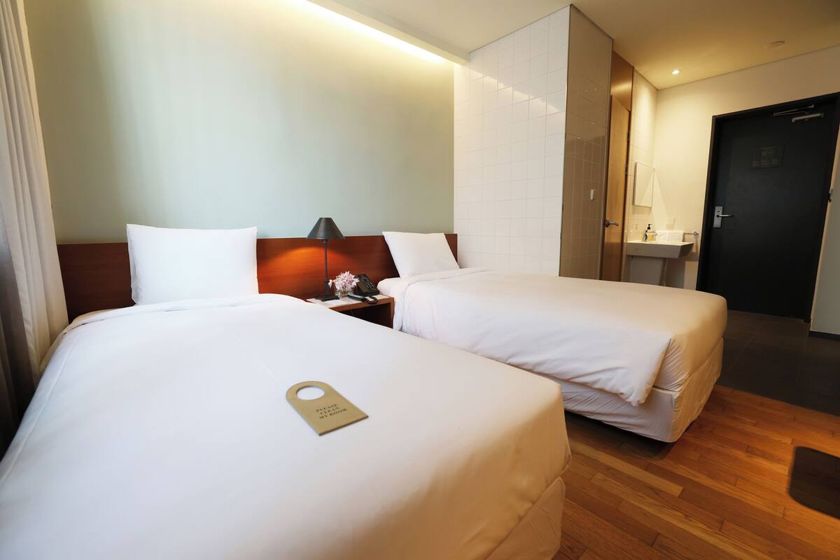 A hotel room with two beds, a bedside table with a lamp, a small table with a phone, and a bathroom with a sink and mirror.