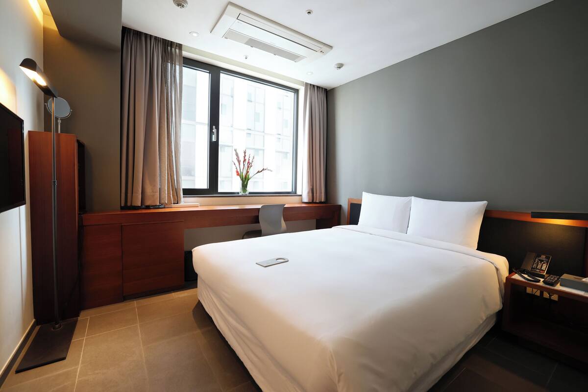 A hotel room with a large bed, a desk, a chair, a nightstand with a phone, and a window with curtains.