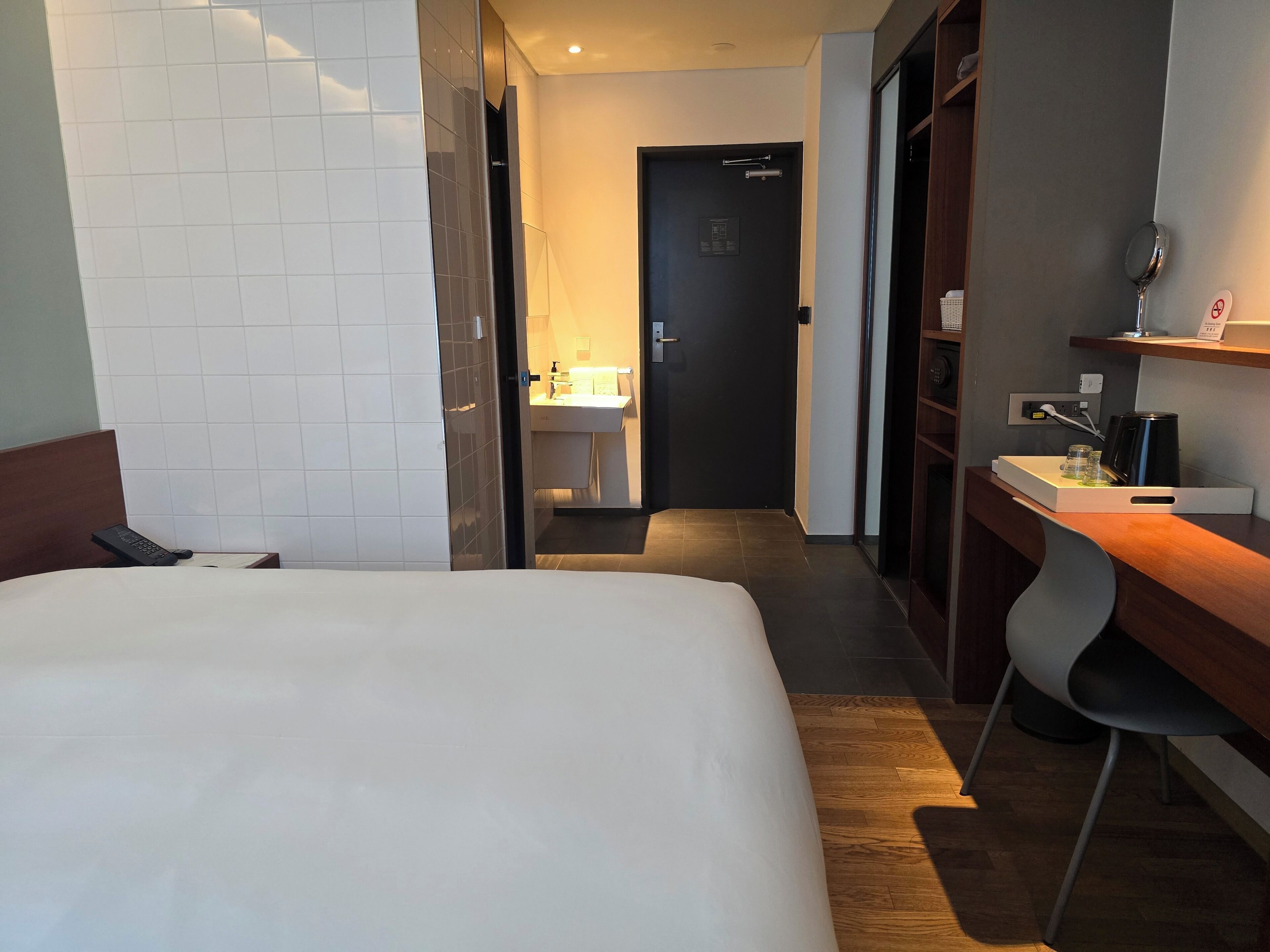A hotel room with a large bed, a desk with a chair, a mirror, and a small bathroom with a sink and towel rack.