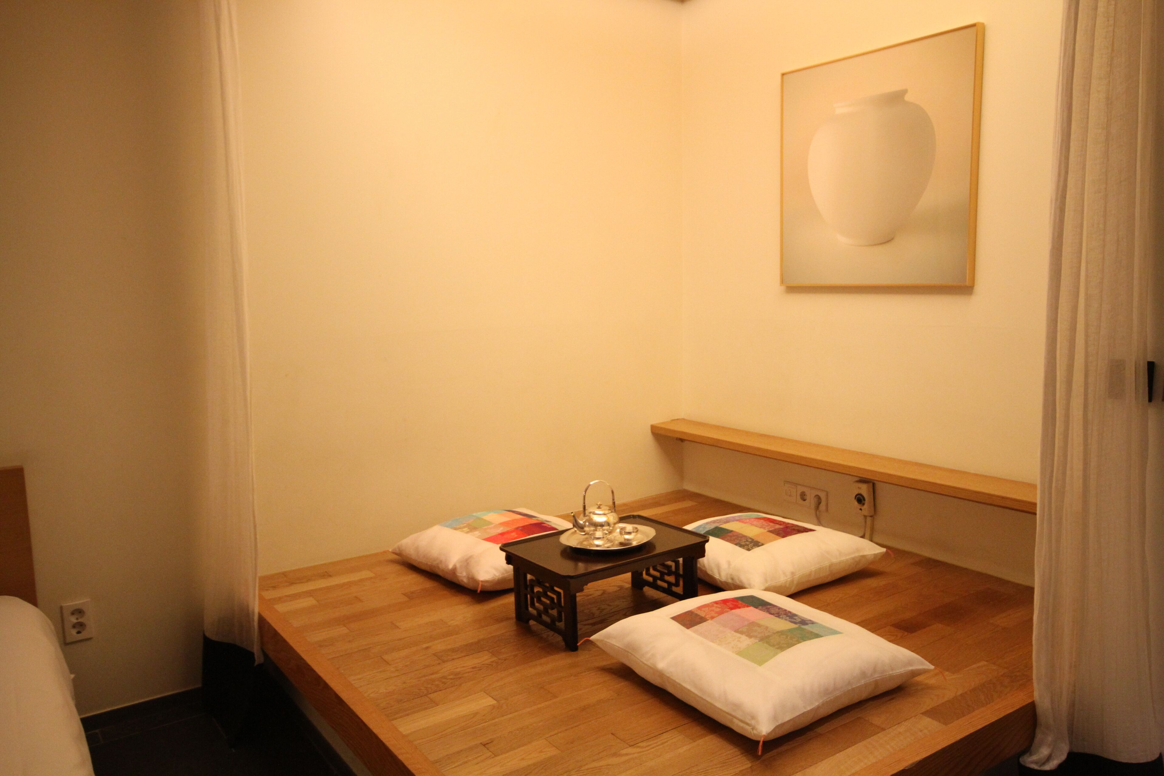 A neatly arranged room with a wooden bed, a small table with a teapot, and a cushion with a colorful patchwork design.
