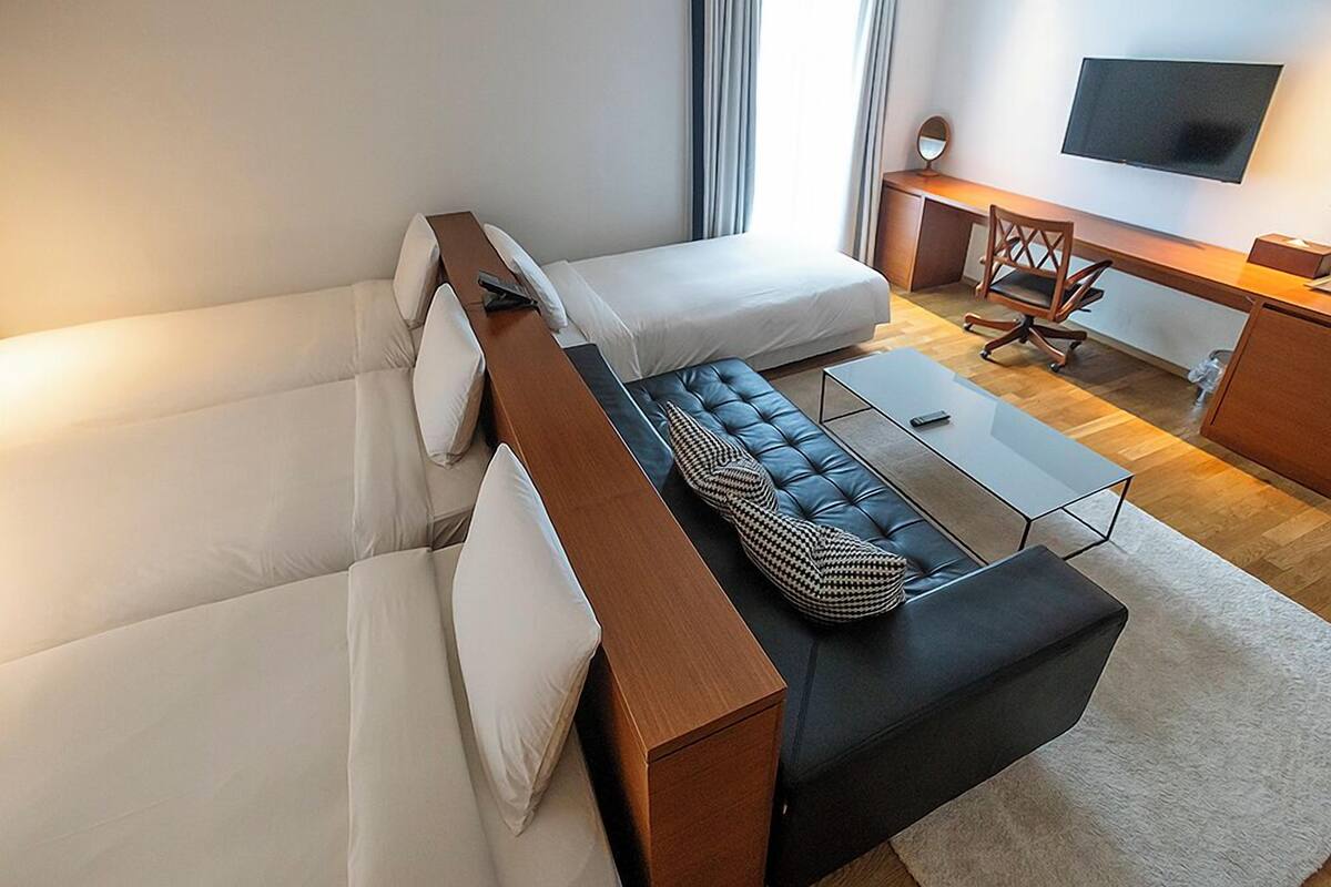 A modern hotel room with a sofa bed, a separate couch, a small table, a desk with a chair, and a flat-screen TV mounted on the wall.