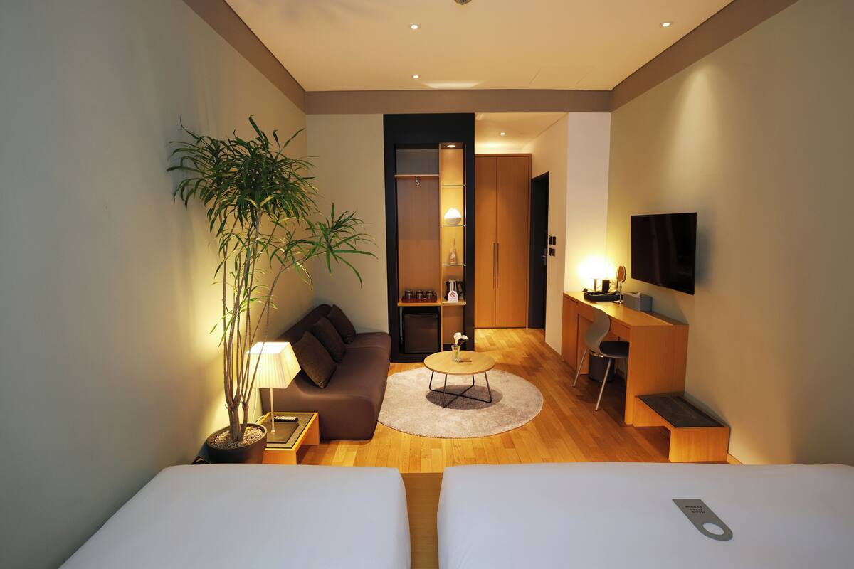 A modern hotel room with two beds, a sofa, a dining table, a TV, and a potted plant.