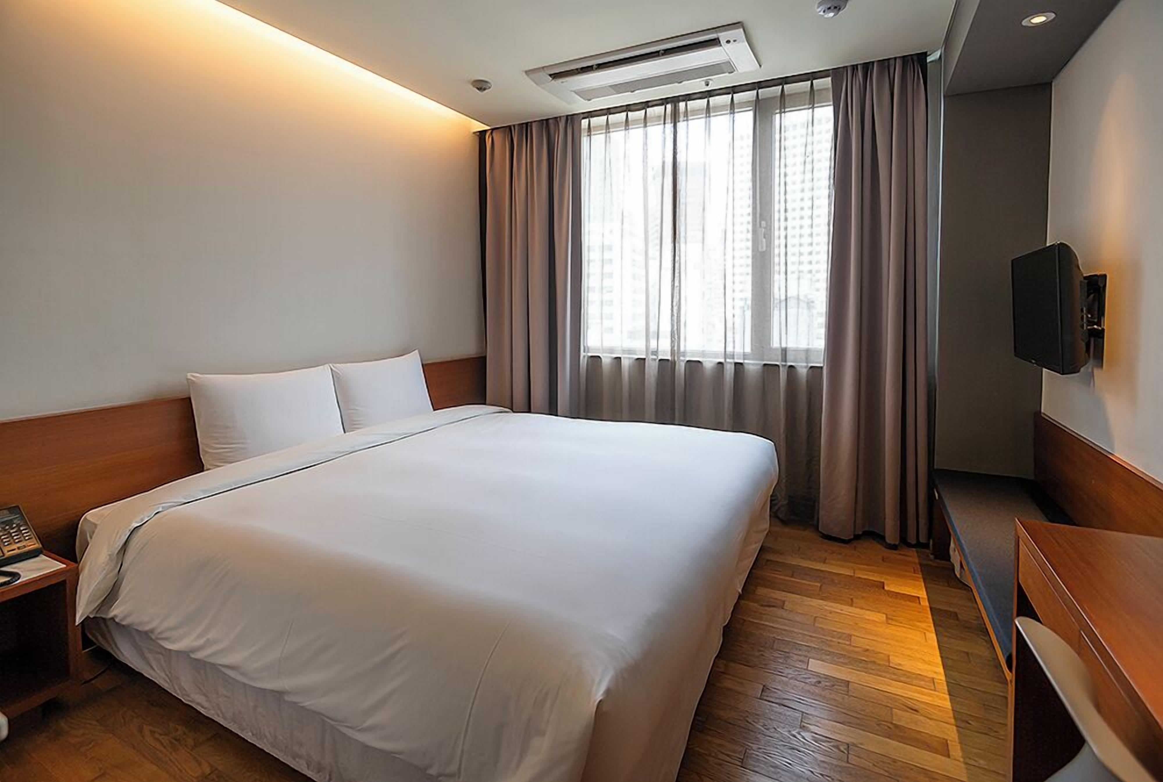 Habitación de hotel con una cama grande, una mesita de noche con lámpara, un televisor montado en la pared y una ventana con cortinas.