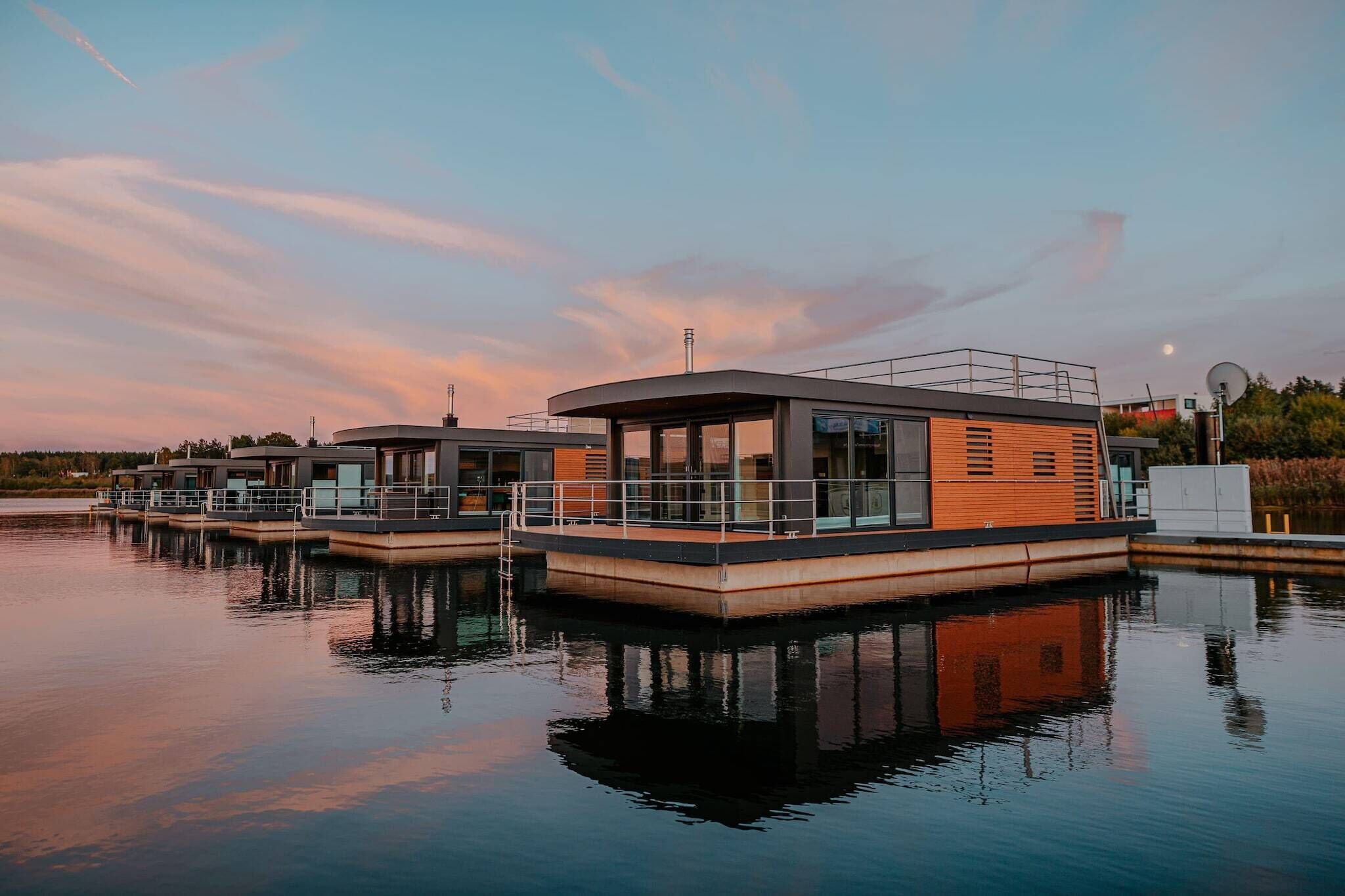 A modern floating structure with a covered deck and glass walls, situated on calm water.
