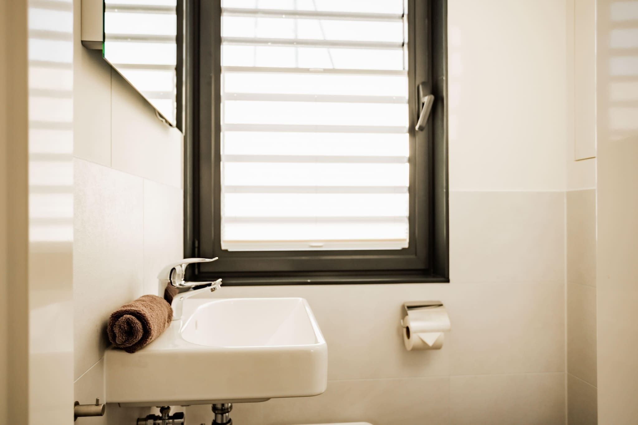 A bathroom with a white sink, a towel, a window with blinds, and a toilet paper holder.