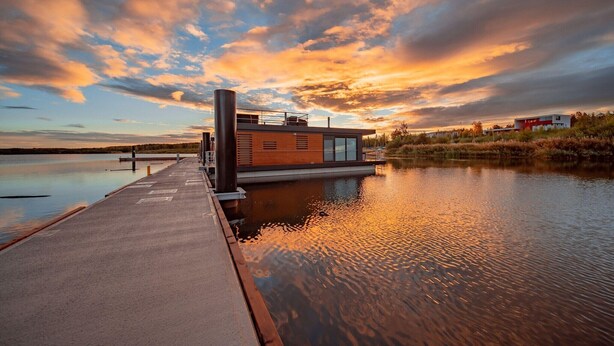 Une péniche moderne, dotée d’une coque en bois et d’une cheminée en métal, est amarrée sur un lac paisible, sous un ciel embrasé par le coucher de soleil.