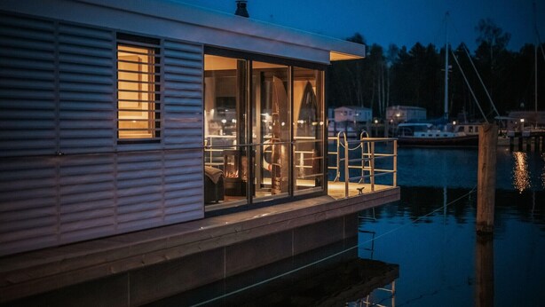 Een woonboot aan het water met een glazen dek, een zichtbaar interieur met een open haard, en een steiger die in het water reikt.