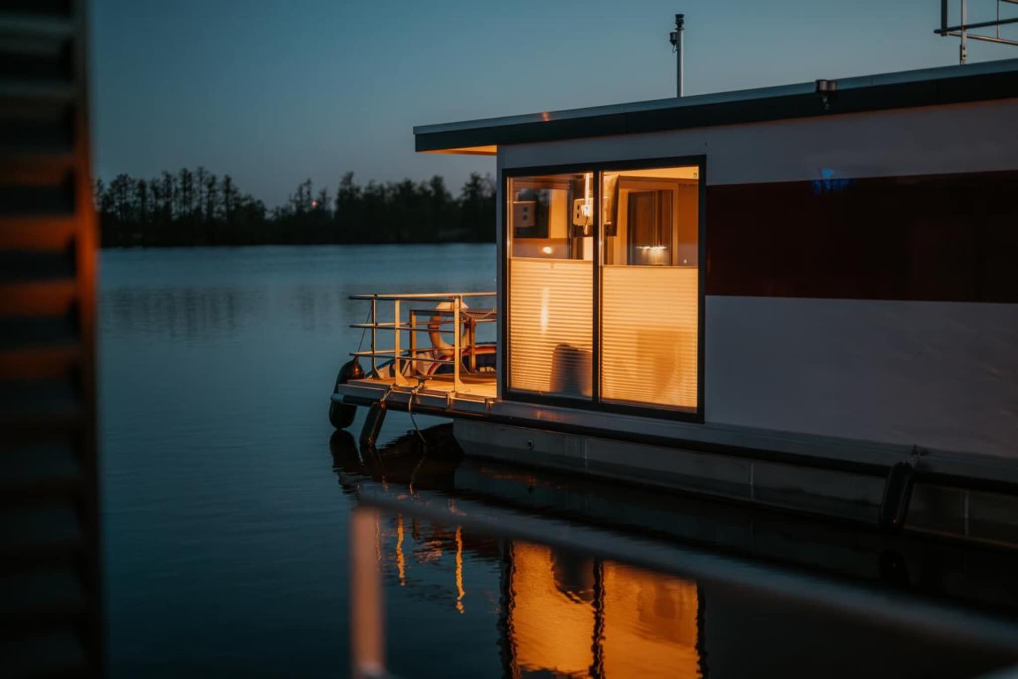Une maison flottante moderne dotée d’une porte en verre, d’un petit espace extérieur aménagé pour s’asseoir et offrant une vue sur l’eau.