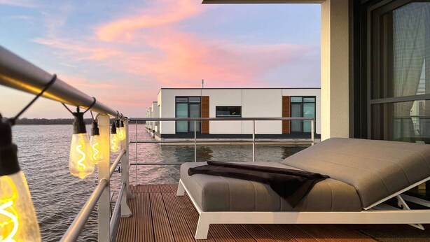 A modern outdoor deck with a lounge area, a view of a modern building across the water, and string lights along the railing. - Bright Base with Coastal Access (Ribnitz-Damgarten)
