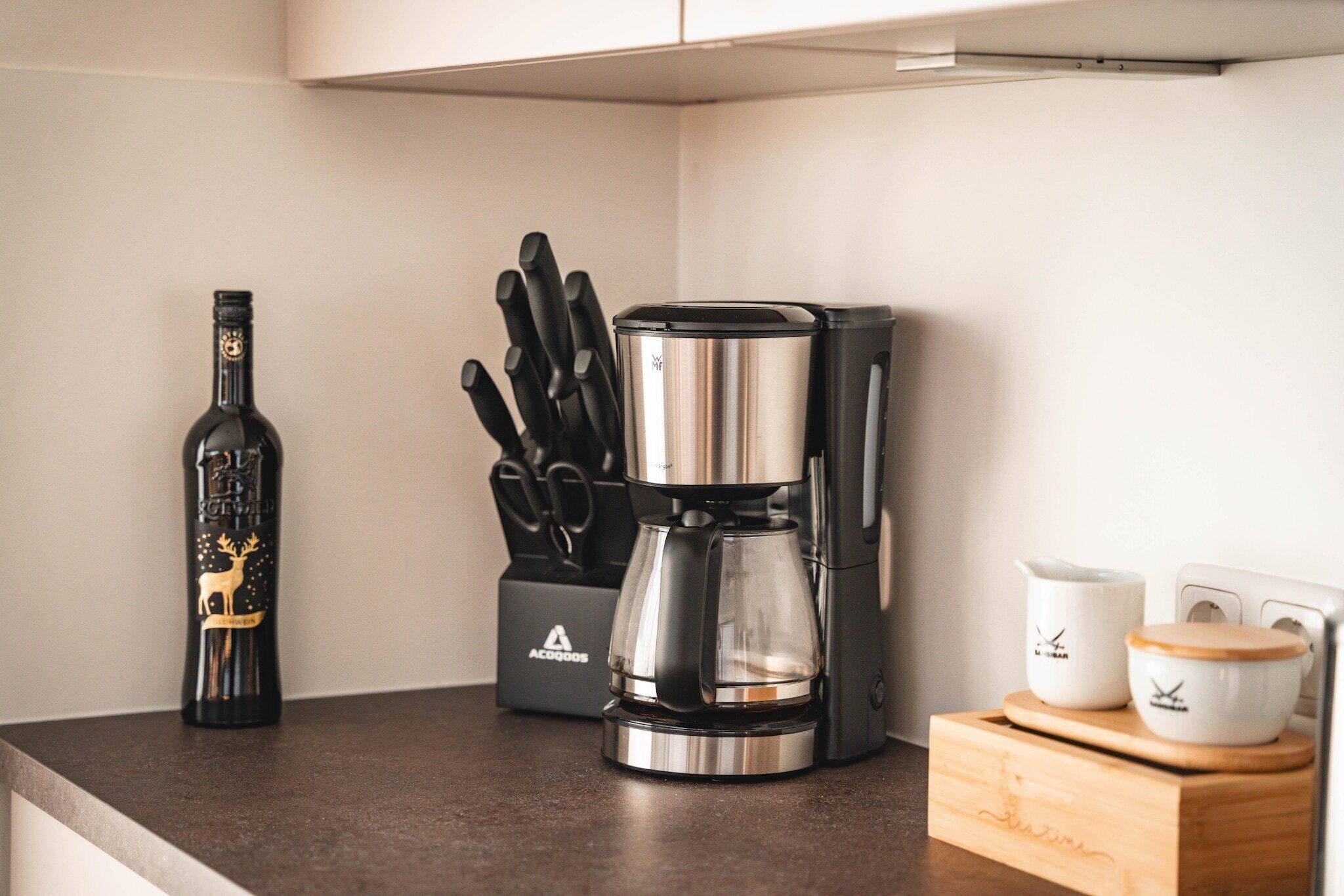 Una encimera de cocina con una cafetera, un bloque de cuchillos, una botella y dos tazas blancas.
