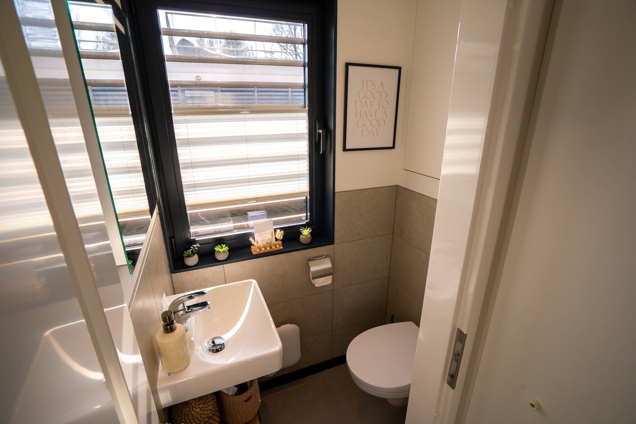 Un bany petit i funcional amb finestra, un lavabo, un vàter i una frase inspiradora emmarcada a la paret.
