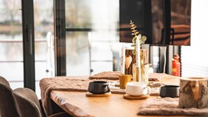 Uma área de refeições acolhedora com uma mesa de madeira posta para dois, com um vaso e uma planta, uma vela e duas chávenas.