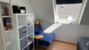 A room with a blue sofa, a small round table, a white shelving unit with books, and a window with a view of a tiled roof. - Sandwall 14, Whg. 6, Sandwallkoje - Holiday apartment with 3 rooms on the Sandwall (Wyk auf Föhr)