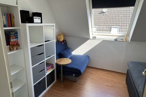Ein Zimmer mit einem blauen Sofa, einem kleinen runden Tisch, einer weißen Bücherregal und einem Fenster mit Blick auf ein Ziegeldach.