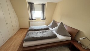 A bedroom with a bed, a nightstand, a lamp, a window with curtains, a radiator, and a wardrobe. - Sandwall 14, Whg. 6, Sandwallkoje - Holiday apartment with 3 rooms on the Sandwall (Wyk auf Föhr)
