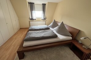 Ein Schlafzimmer mit Bett, Nachttisch, Lampe, einem Fenster mit Vorhängen, einer Heizung und einem Kleiderschrank.