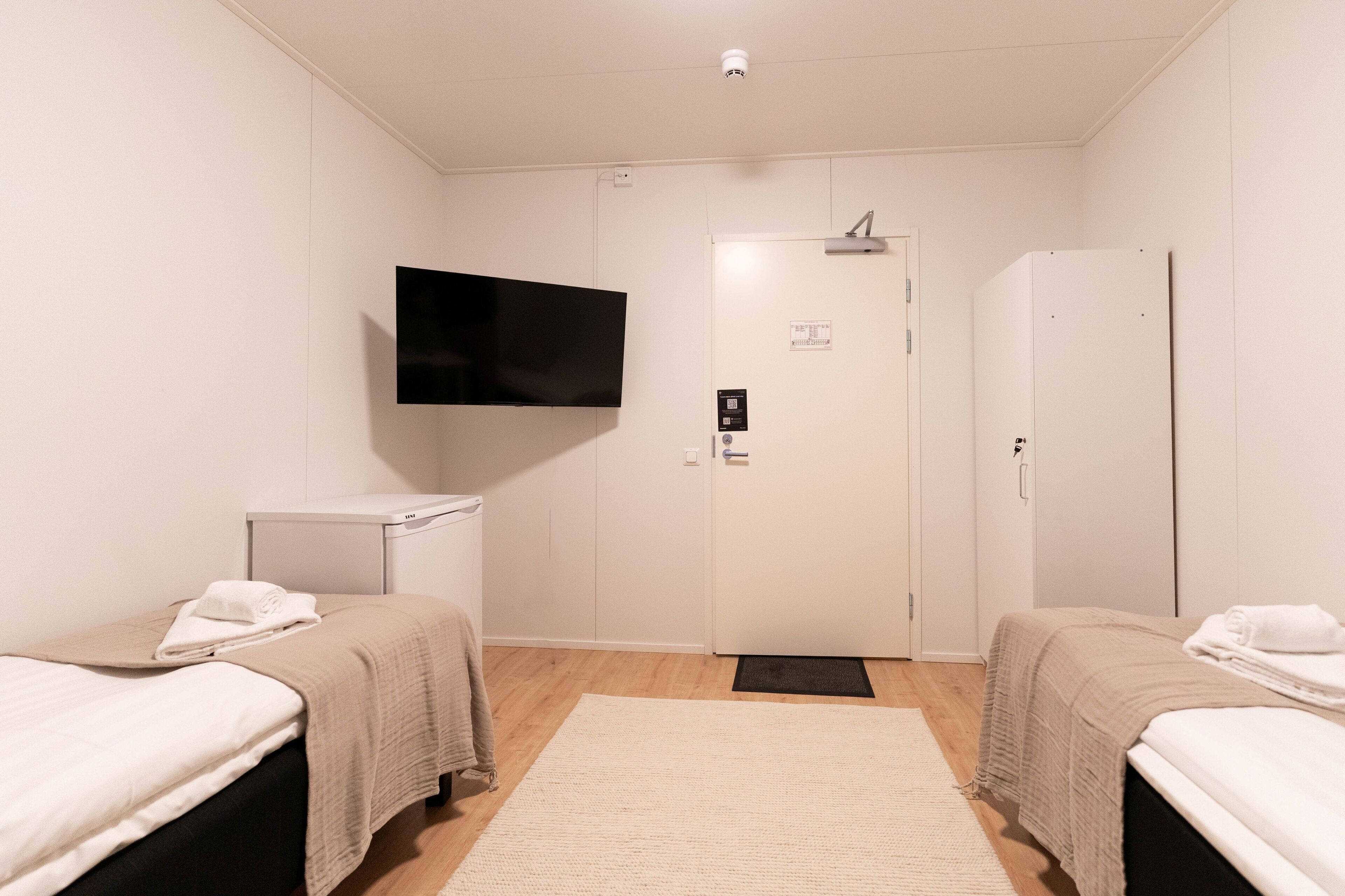 Habitación de hotel con dos camas, un televisor montado en la pared, un armario blanco y una puerta con picaporte.