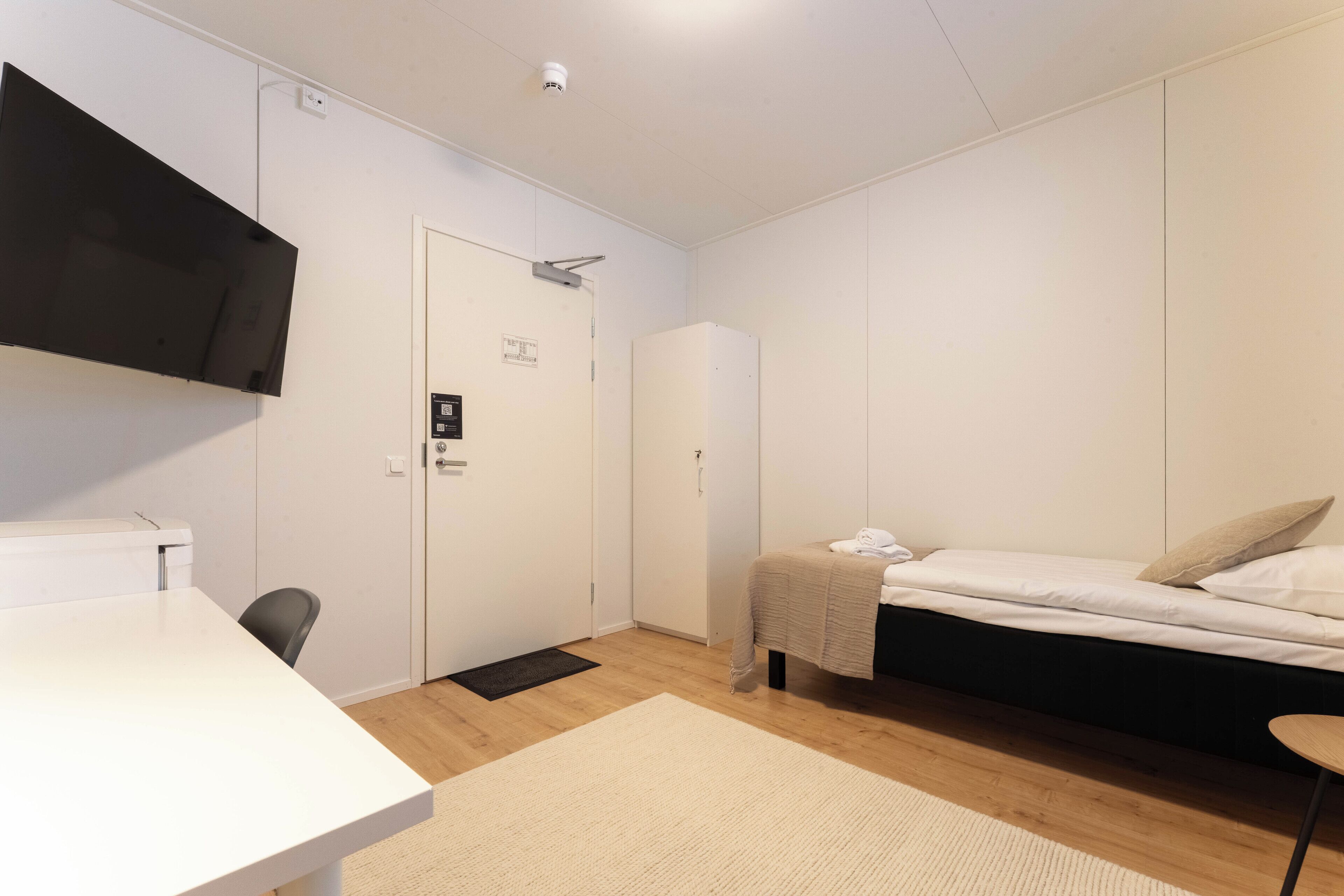 A room with a bed, a desk, a chair, a television mounted on the wall, and a door with a sign.
