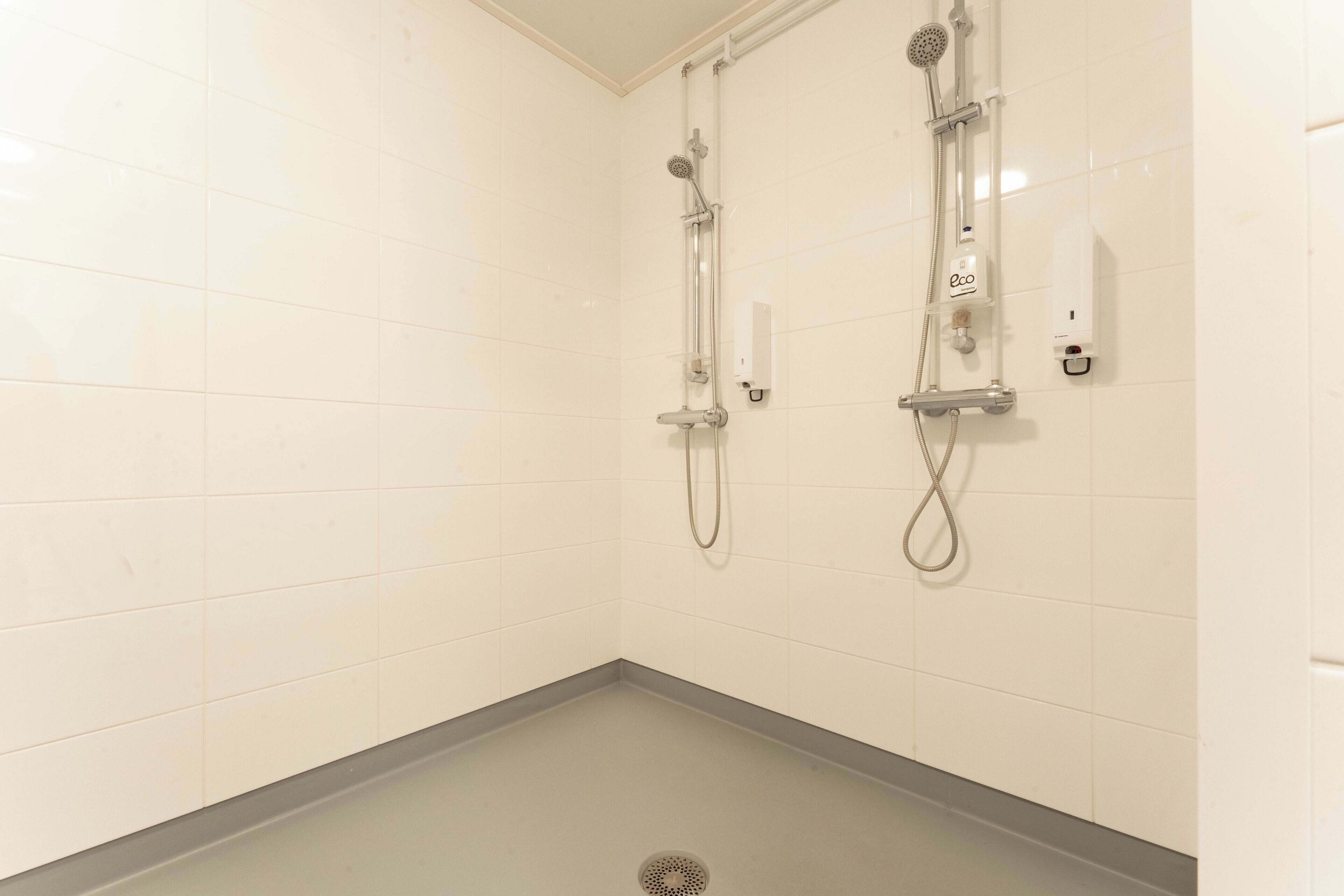 Ein Badezimmer mit weiß gefliesten Wänden und einem grauen Boden. Zwei Duschköpfe sind an der Wand angebracht, und ein Handtrockner ist auf der rechten Seite sichtbar.