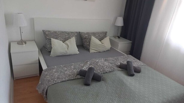 Una cama bien hecha con una colcha estampada y almohadas, dos mesitas de noche con lámparas, y una ventana con cortinas.