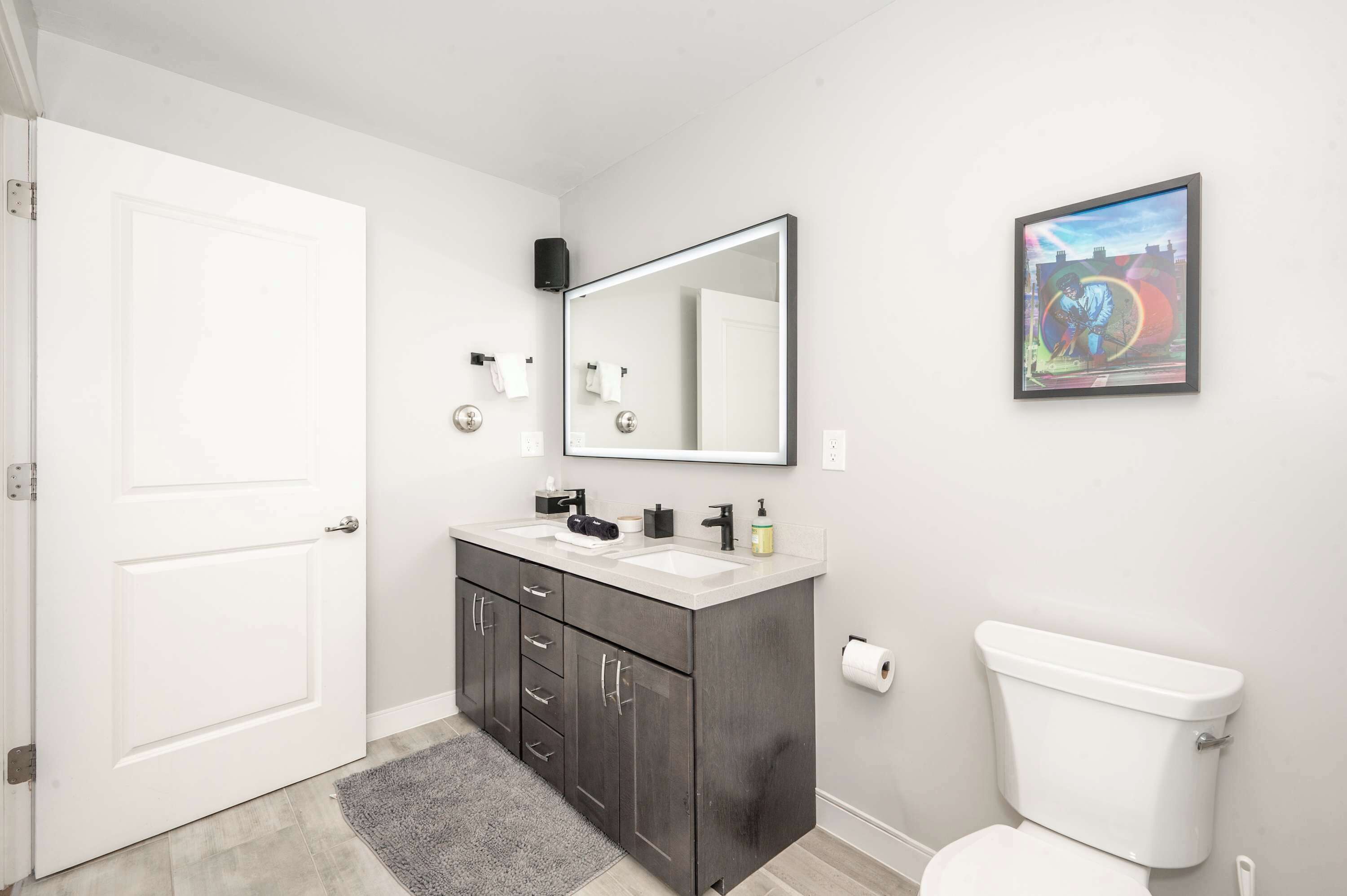 Une salle de bain moderne avec des toilettes blanches, des meubles sombres, un grand miroir et une œuvre d’art encadrée accrochée au mur.