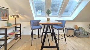 A small, modern room with a sloped ceiling and skylights, featuring a wooden table, two chairs, a small plant, and a shelf with books and decorative items. - Apartment Turmblick - Comfortable apartment in the center of Marienhafe with (Marienhafe)