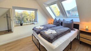 A bedroom with a skylight, a bed with gray bedding, a bedside table with a lamp, and a window with a view of trees. - Apartment Turmblick - Comfortable apartment in the center of Marienhafe with (Marienhafe)