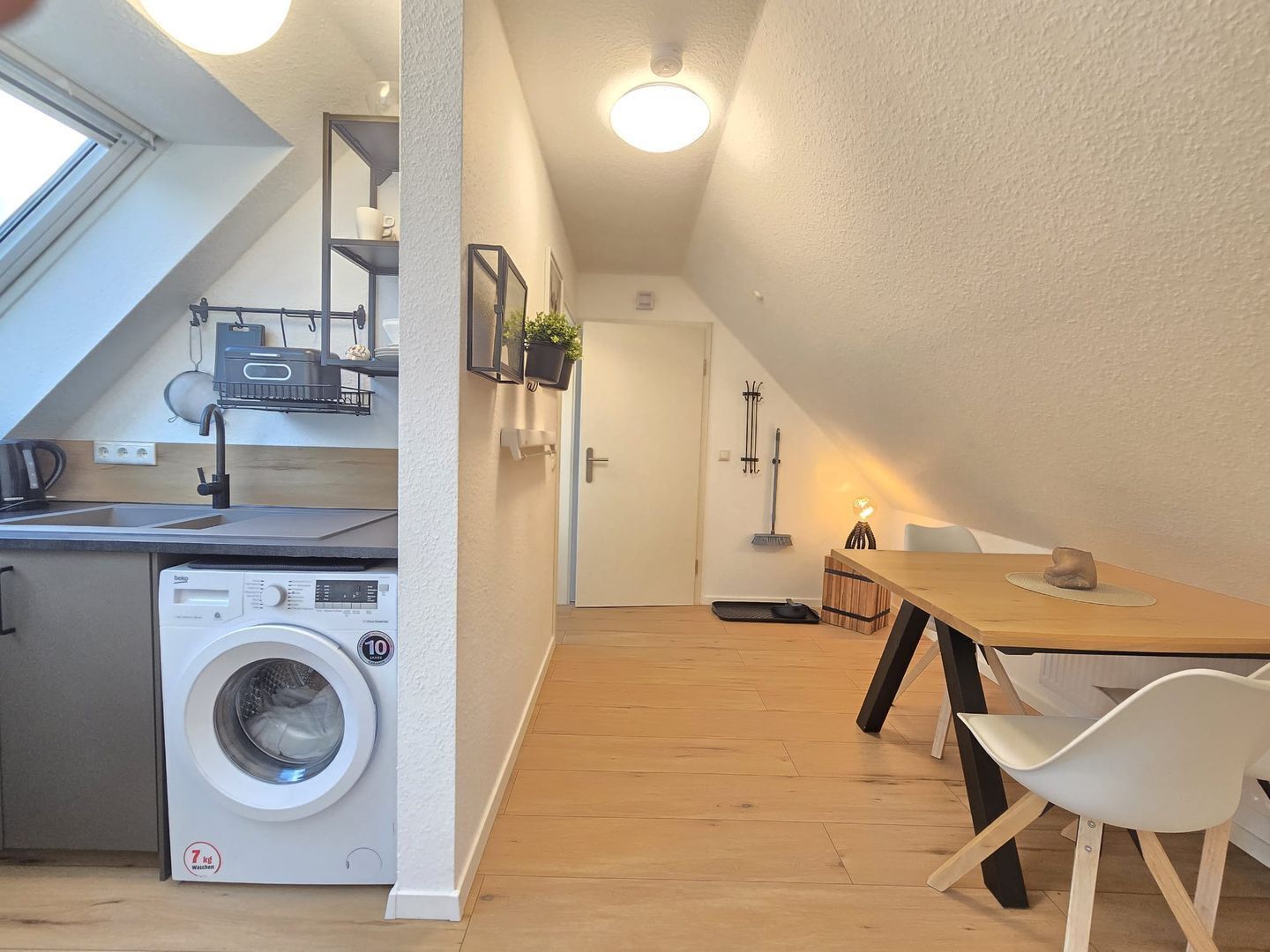 A compact kitchen with a built-in washer, a dining area with a wooden table and white chairs, and a small hallway with a door and a broom.