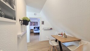 Un pasillo estrecho con suelo de madera, paredes blancas y una pequeña zona de comedor con una mesa y dos sillas. Se ve una cama al fondo, y una planta cuelga de la pared.