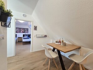 Ein kleiner, moderner Raum mit einem Holztisch und weißen Stühlen, einem Bett mit blauer Bettwäsche sowie einer Wanduhr.