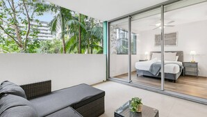 Een balkon met een bank en een kleintje tafeltje, een slaapkamer met een bed en nachtkastjes, en uitzicht op palmbomen en gebouwen.