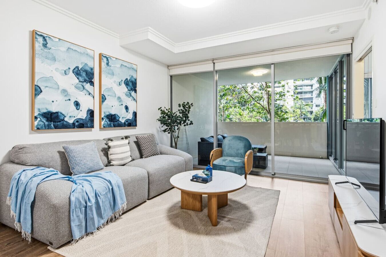 A modern living room with a grey sofa, blue cushions, a round coffee table, and a blue armchair. There are two abstract paintings on the wall, a plant, and a sliding glass door leading to an outdoor balcony.