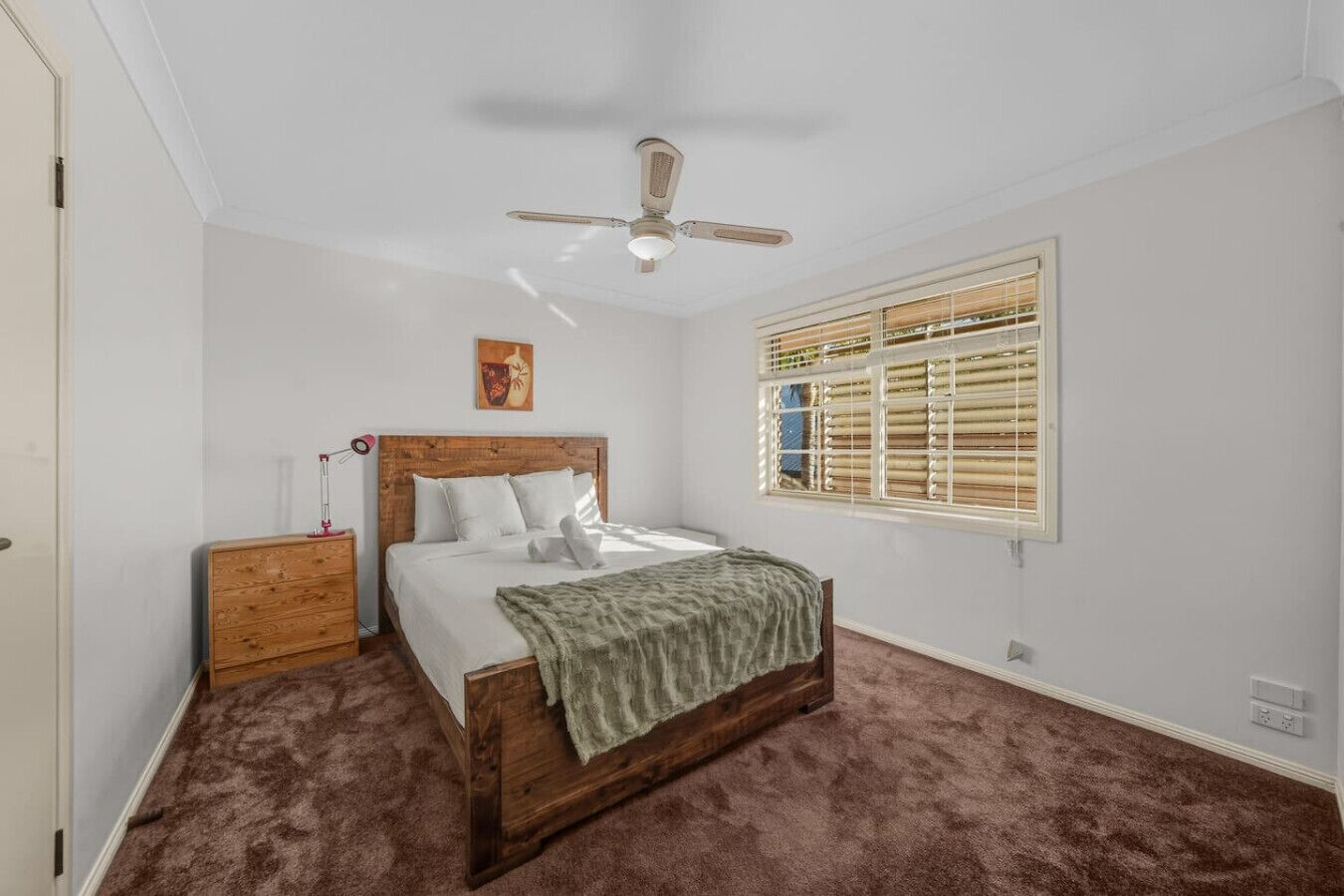 A bedroom with a wooden bed, a ceiling fan, a window with blinds, and a small bedside table with a lamp.