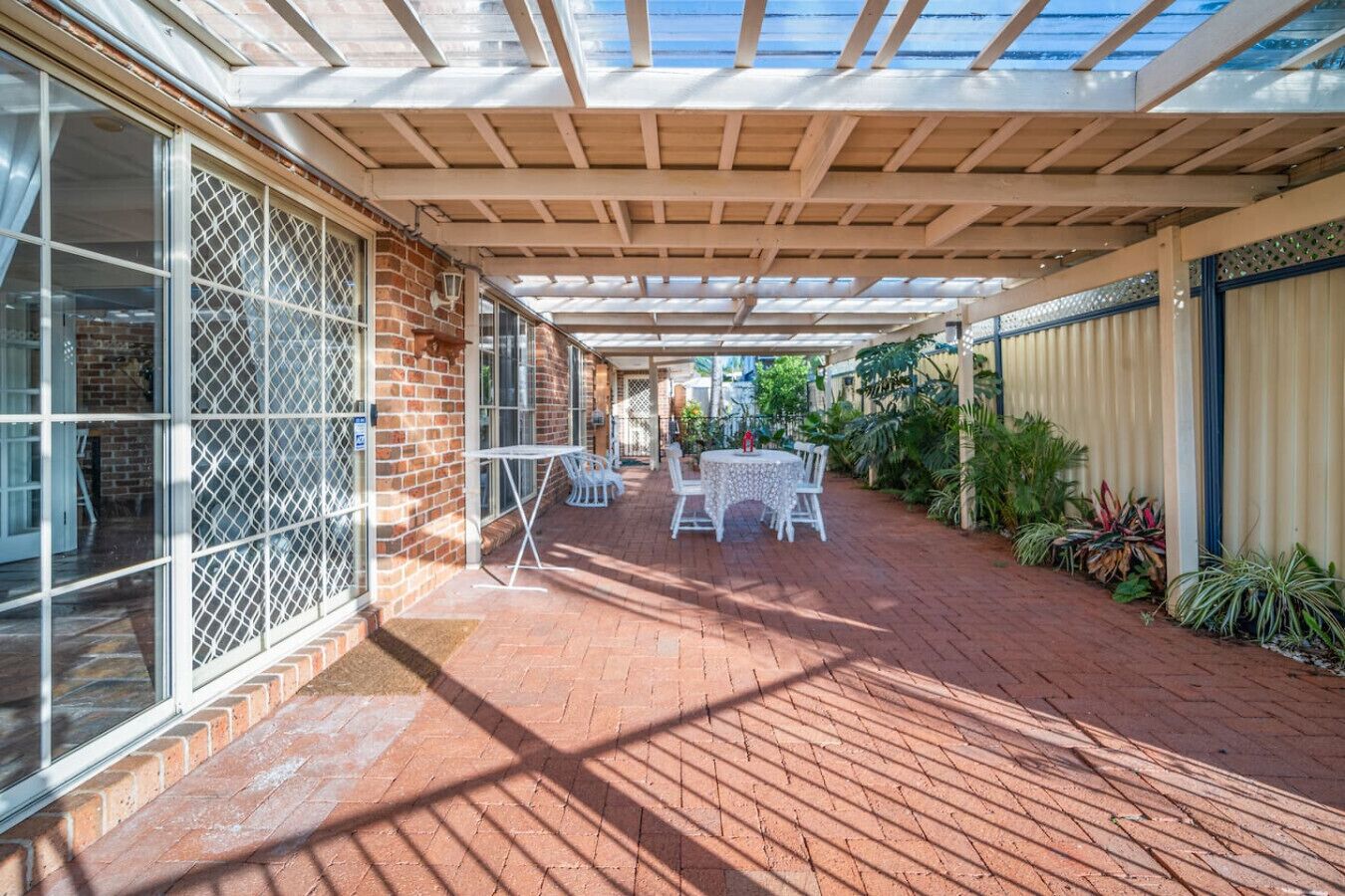 A covered patio with a brick floor, white outdoor furniture, and a glass door on the left.