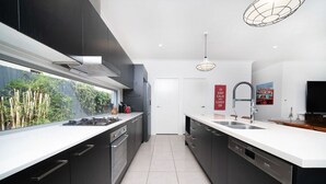 Une cuisine moderne avec des meubles de couleur sombre, un plan de travail blanc, une cuisinière à gaz, un évier et une grande fenêtre donnant sur un espace verdoyant.