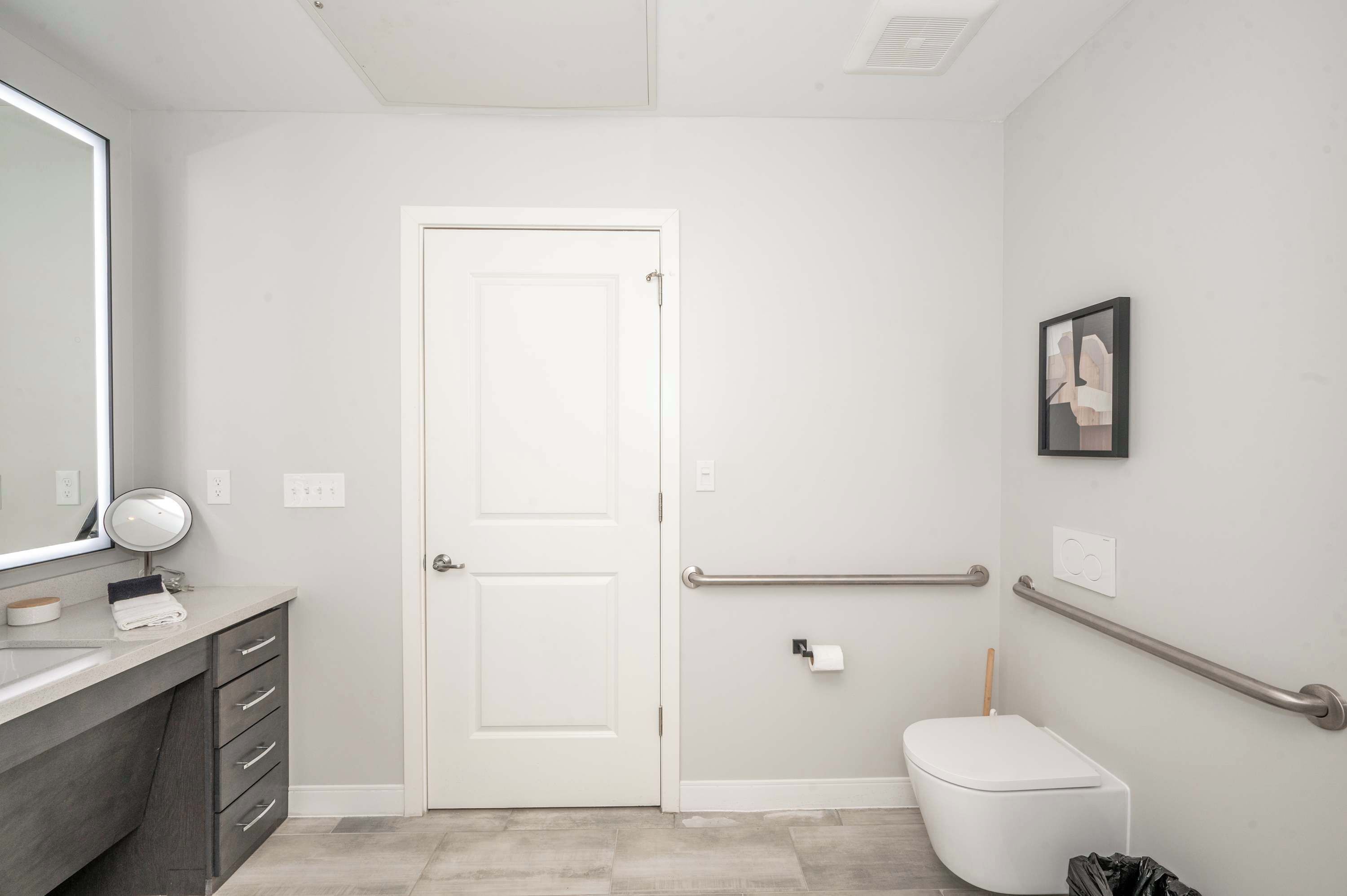 A modern bathroom with a white door, a toilet with a grab bar, a mirror, and a towel rack.
